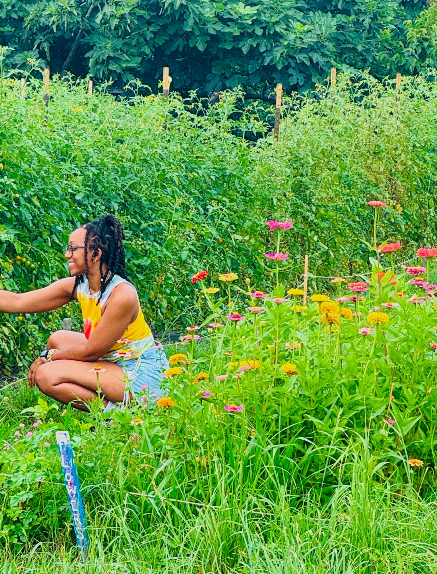 Person sitting cross-legged in a garden with tall green plants and colorful flowers, surrounded by lush greenery.