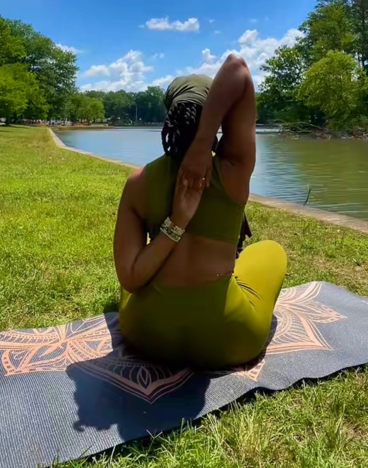 Person practicing yoga outdoors on a mat near a lake, with trees and blue sky in the background.