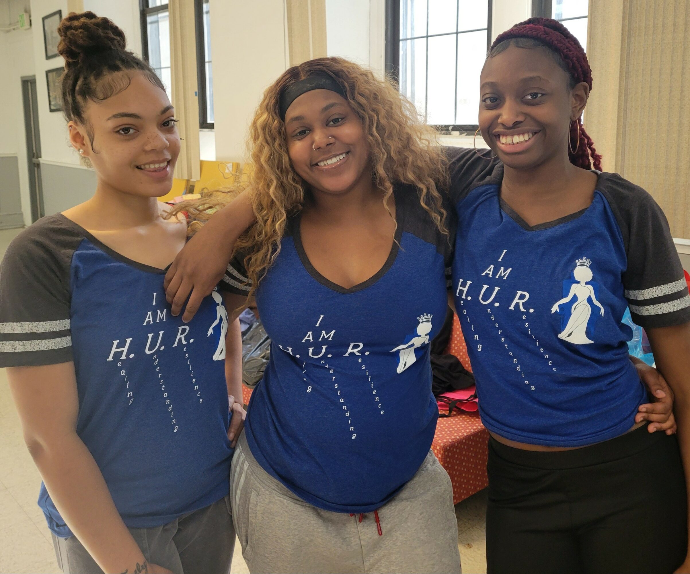Three women wearing matching blue T-shirts with white text and graphics, smiling indoors with large windows in the background.