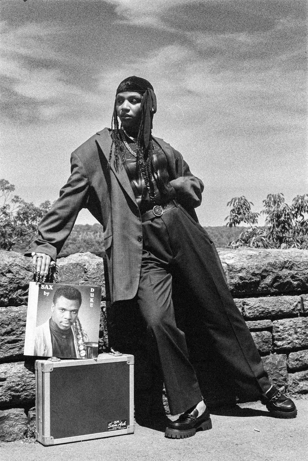 Person with long hair and a blazer standing outdoors next to a small table with a photo and a box, against a cloudy sky.