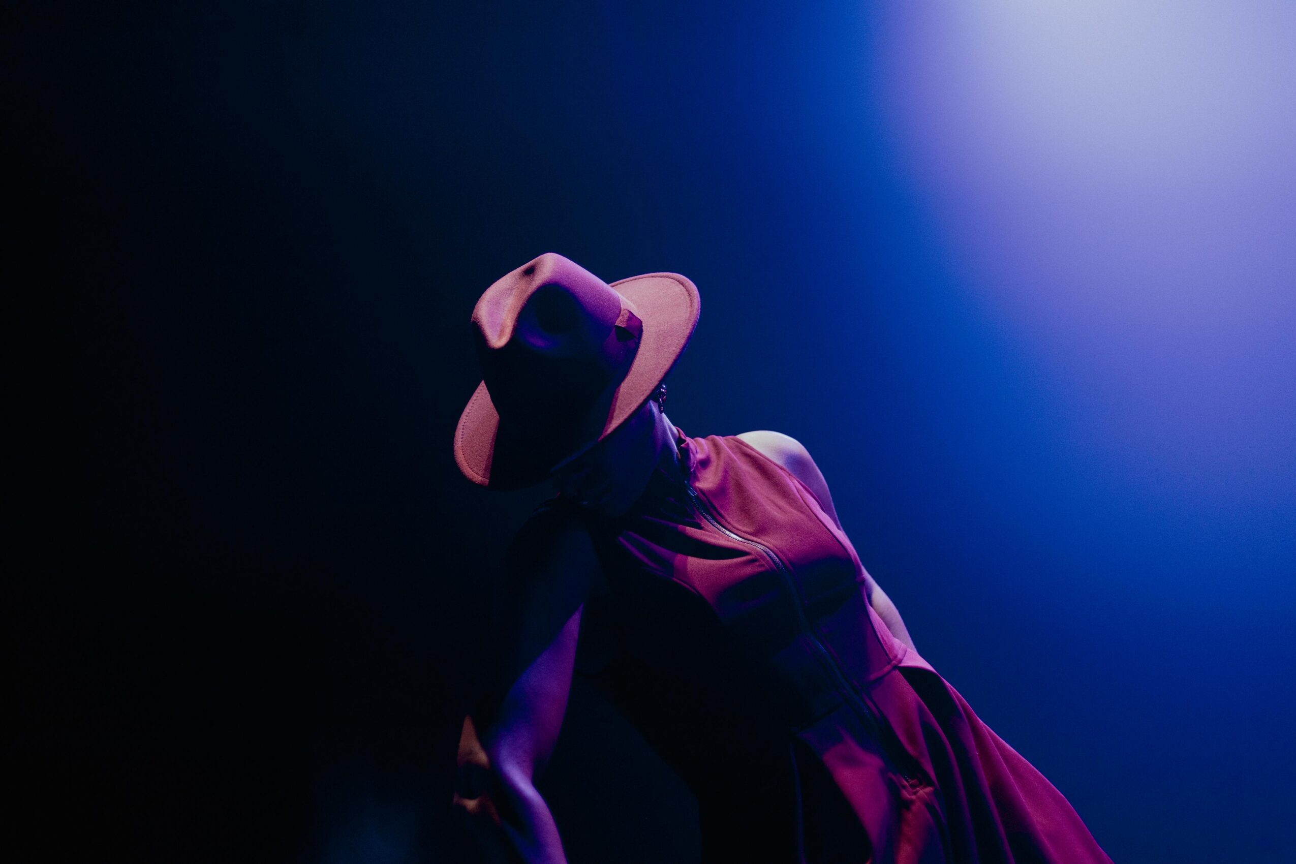 Person wearing a hat and dress, illuminated by blue and purple lighting, standing in a dark environment.