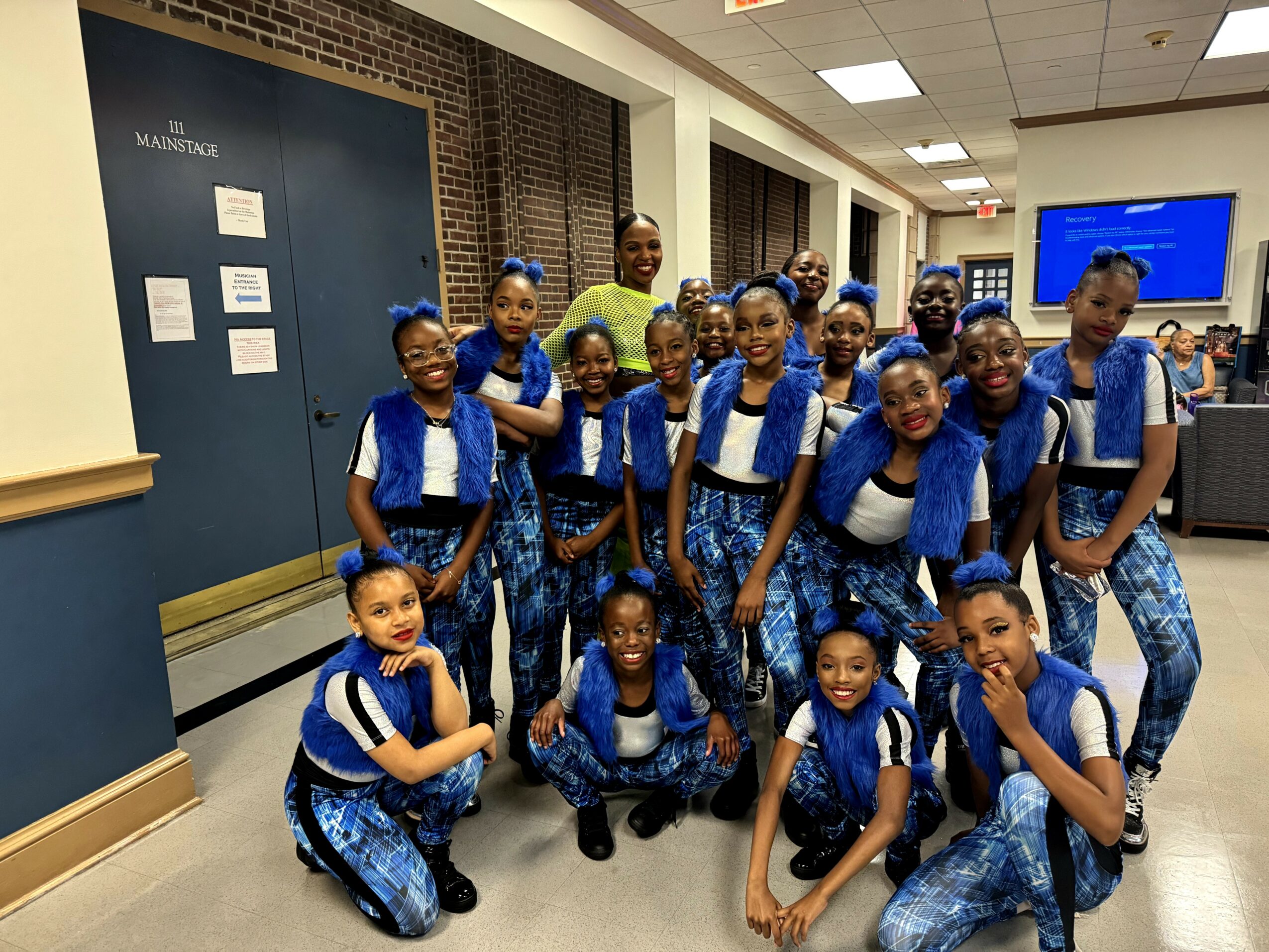 Group of young girls in matching blue and white outfits posing indoors, some kneeling and some standing, smiling at the camera.