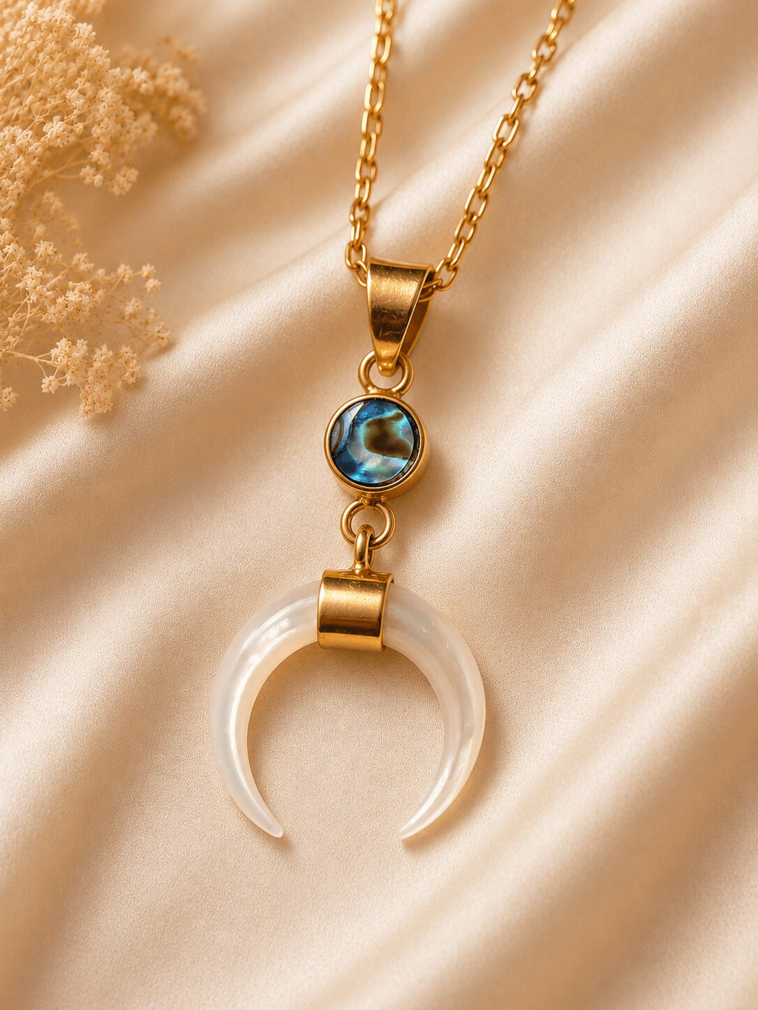 Gold chain necklace with a circular blue gemstone and a crescent-shaped pendant, resting on satin fabric.