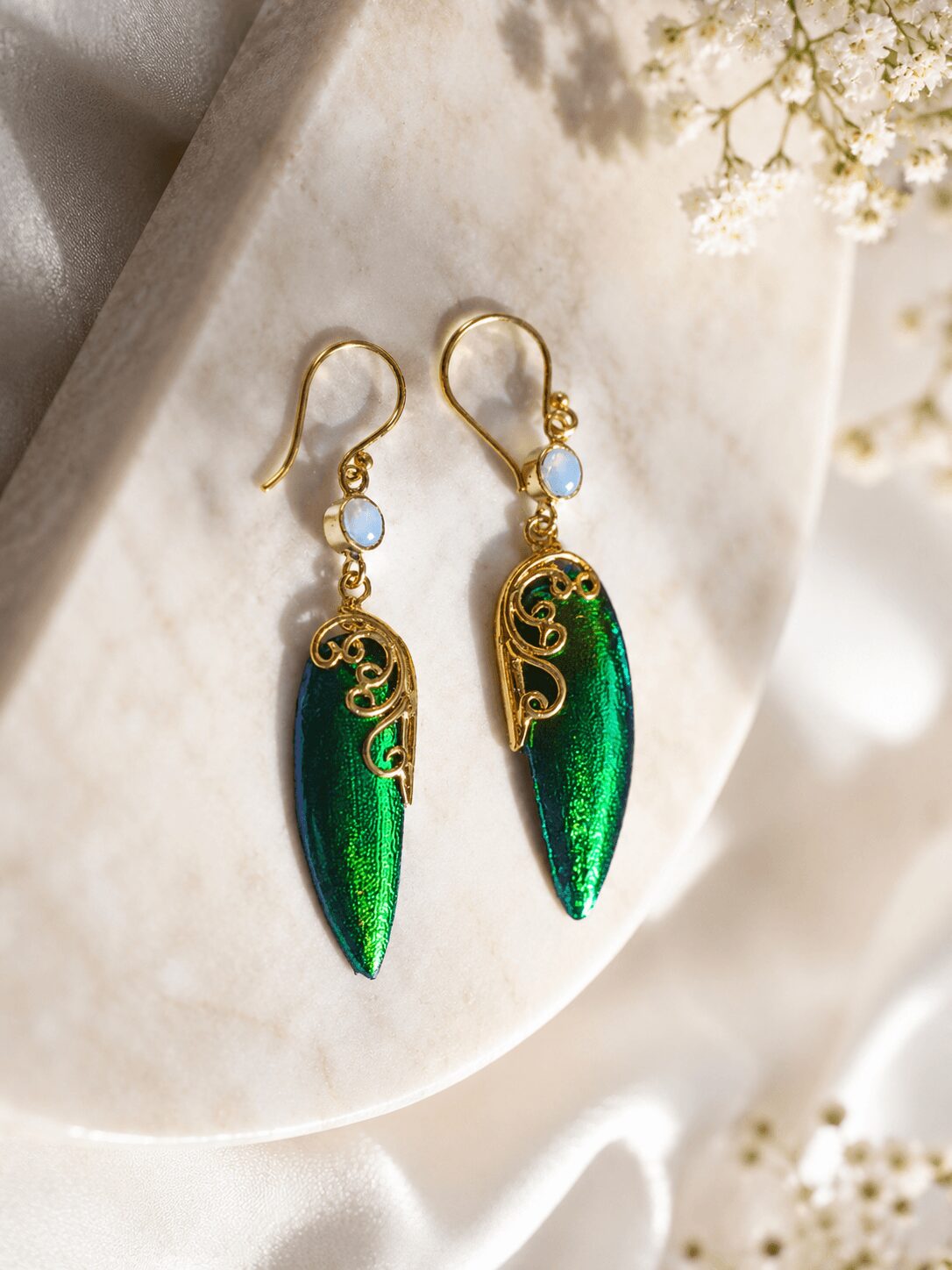 Pair of gold earrings with green and blue elongated pendants, small blue beads, and intricate gold scrollwork, on a white surface.