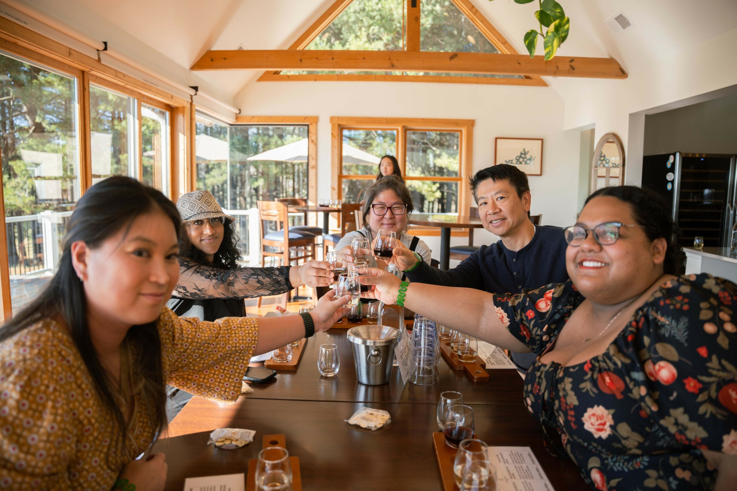 Group of six people sitting at a dining table, raising glasses in a toast, in a bright room with large windows and wooden beams.