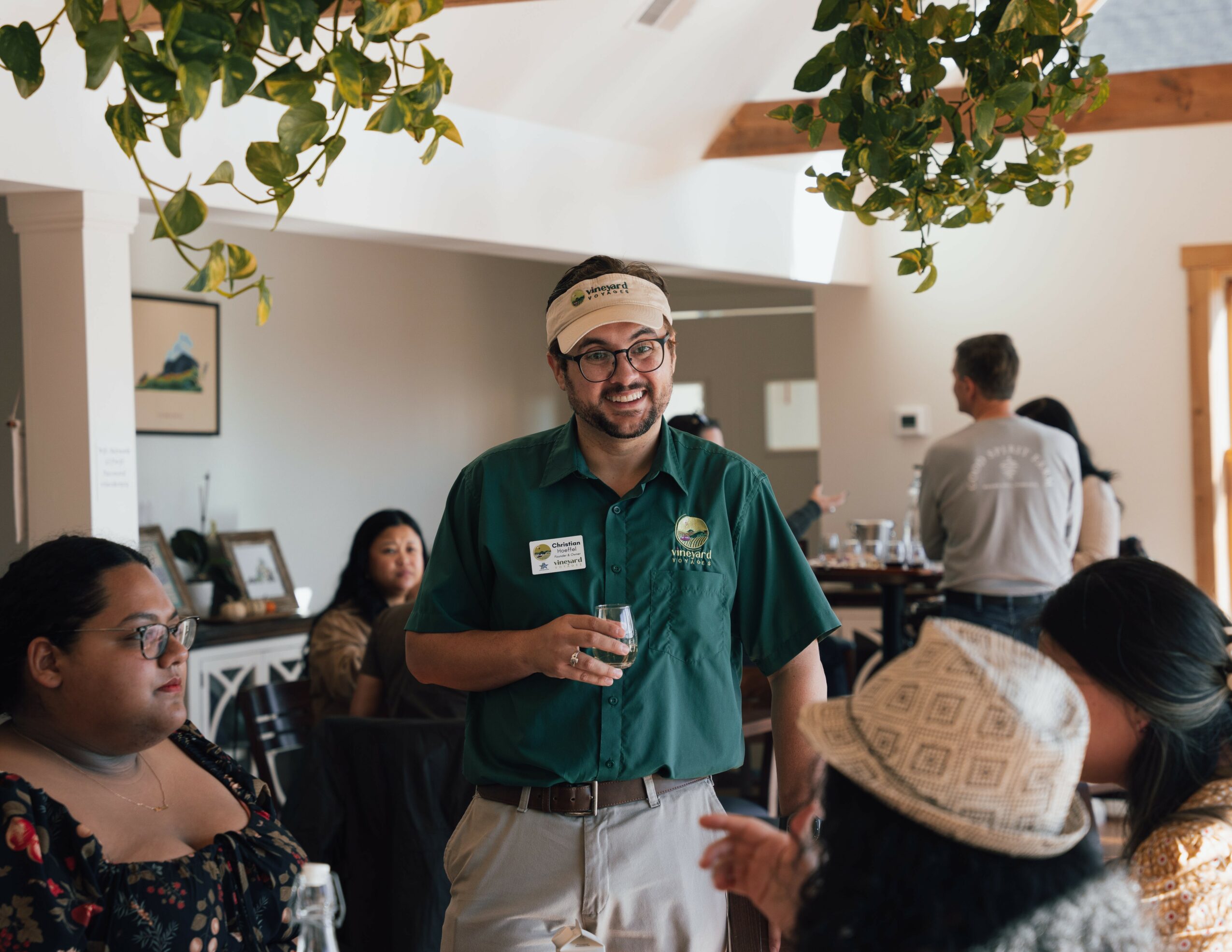 Man with glasses and beard wearing a green shirt and name tag, holding a glass, speaking to a group indoors.