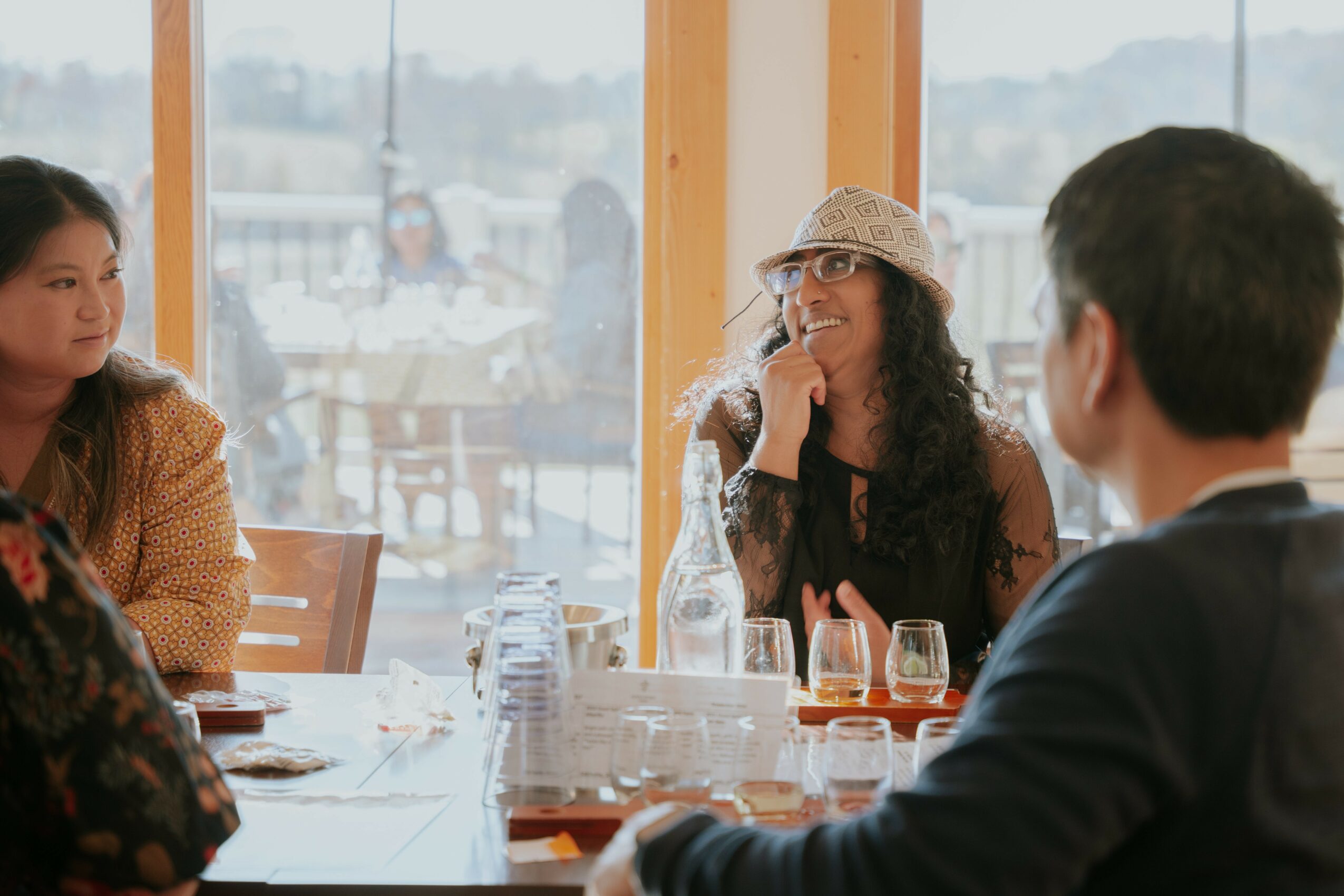 Group of people sitting at a table in a bright room with large windows, engaged in conversation, drinks on the table.