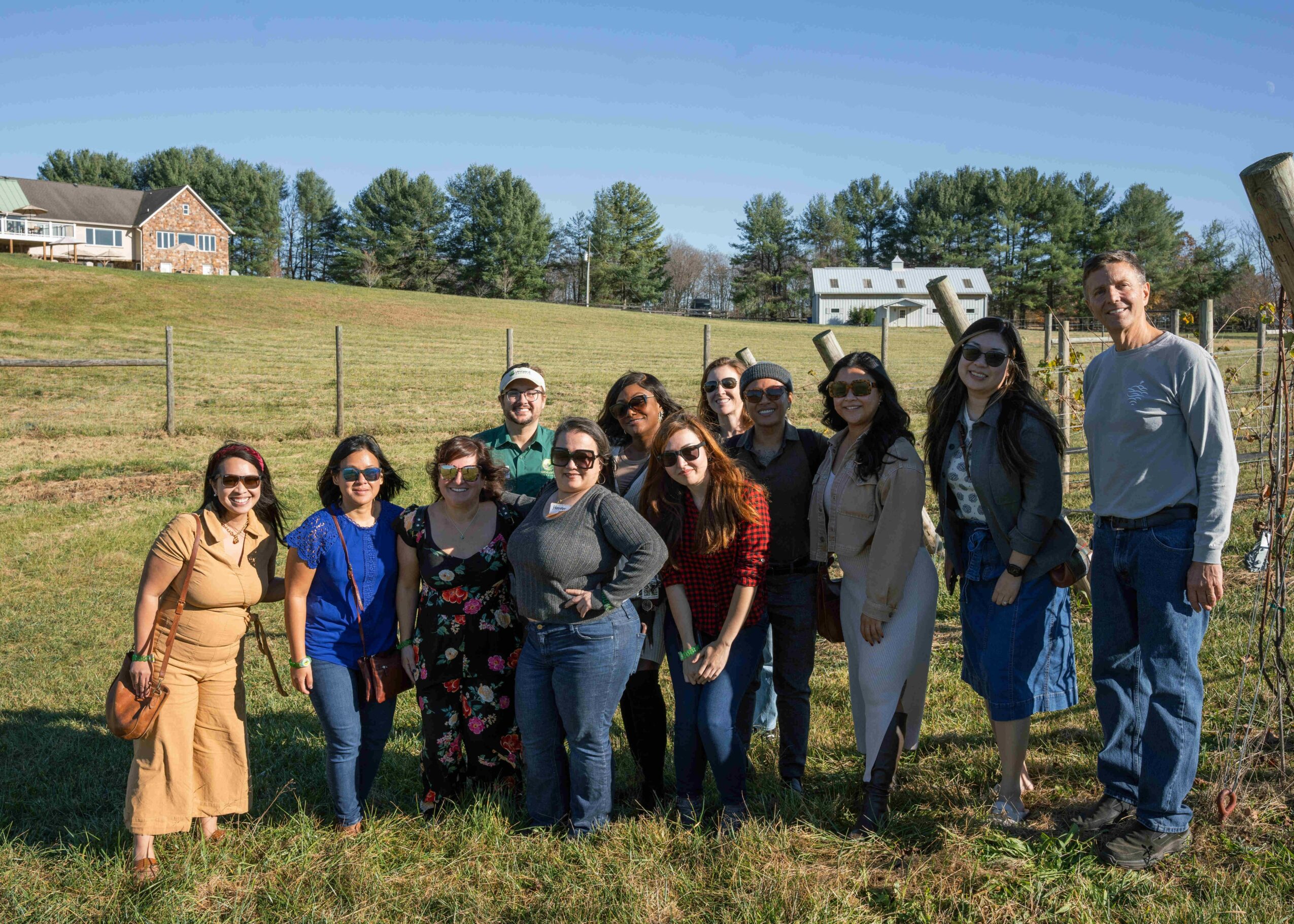 Group of twelve people standing outdoors on grass, with houses and trees in the background, sunny day.