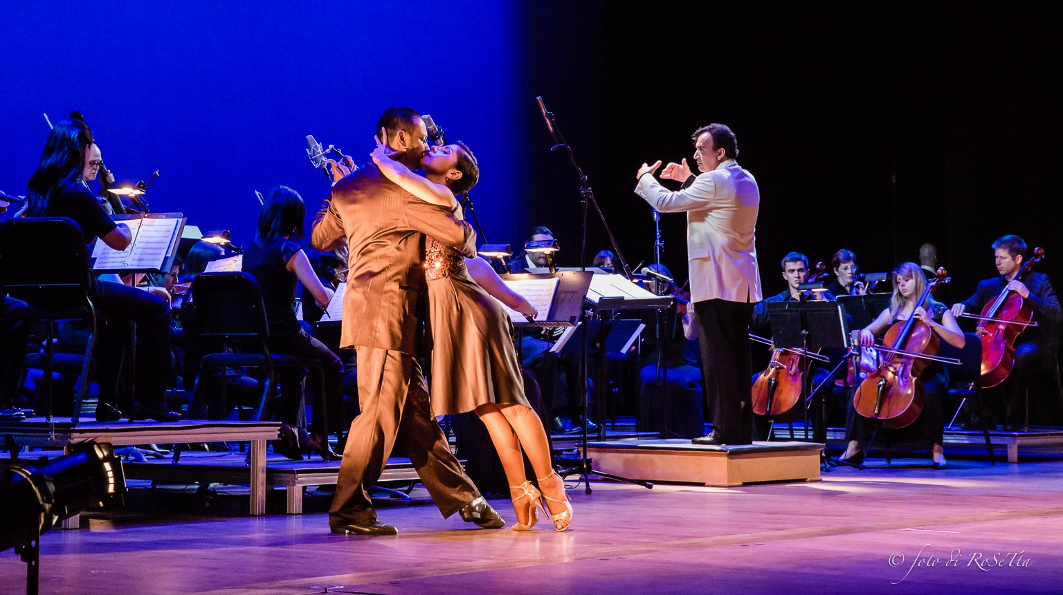 Two dancers perform on stage with an orchestra in the background, one dancer lifts the other in a dance pose.