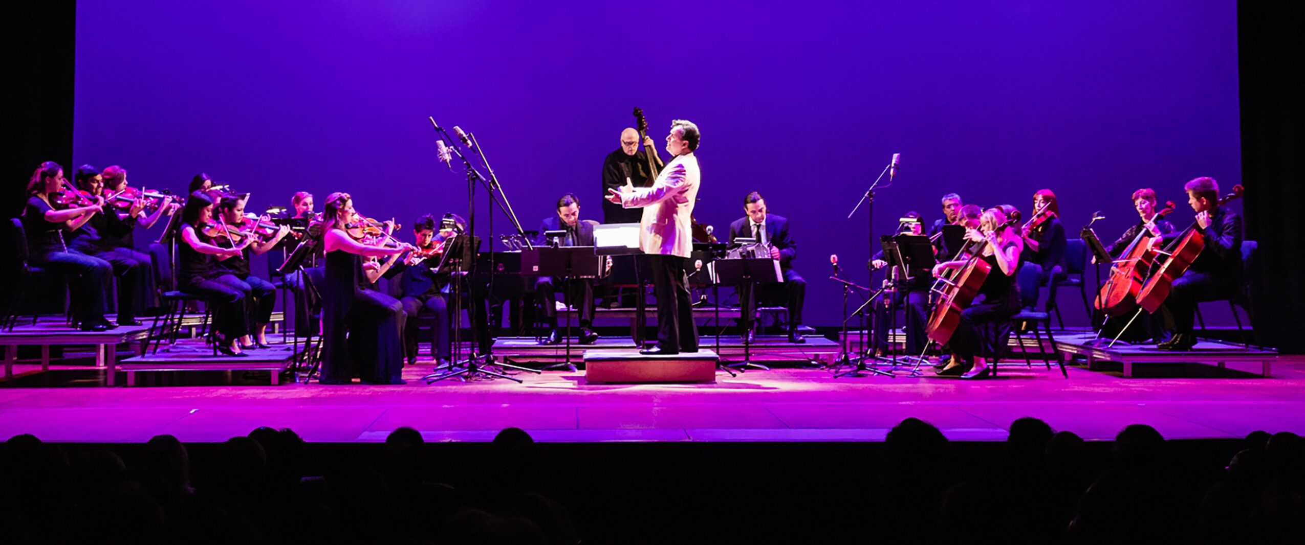 Orchestra performing on stage with conductor and soloist, purple background, musicians playing string instruments, stage lighting.