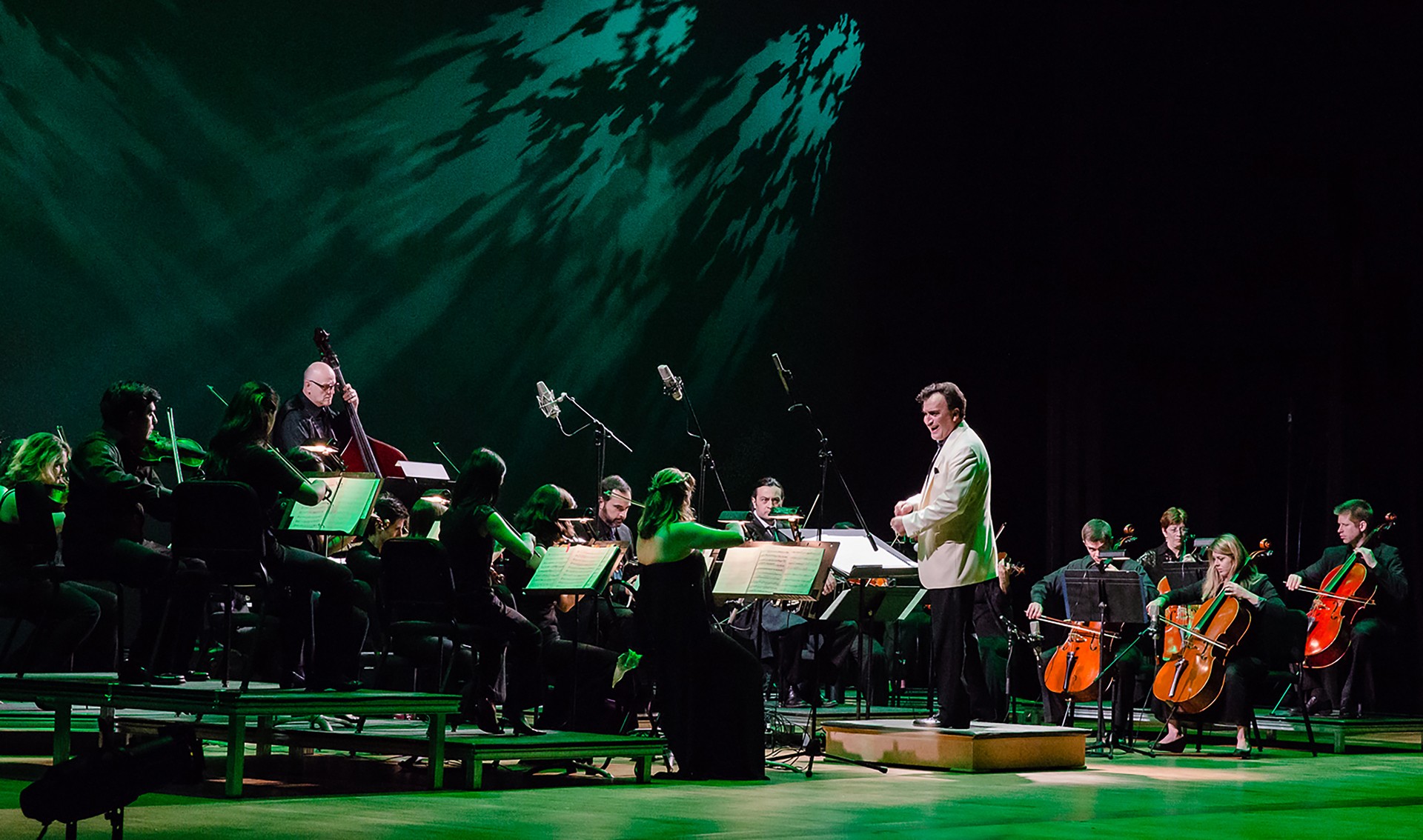 Orchestra performing on stage with conductor, musicians playing string and wind instruments, green lighting effects, dark background.