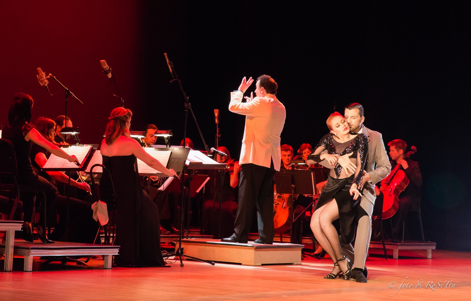 Orchestra performs on stage with conductor and two dancers, stage lit with warm lighting, black background, musicians playing instruments.