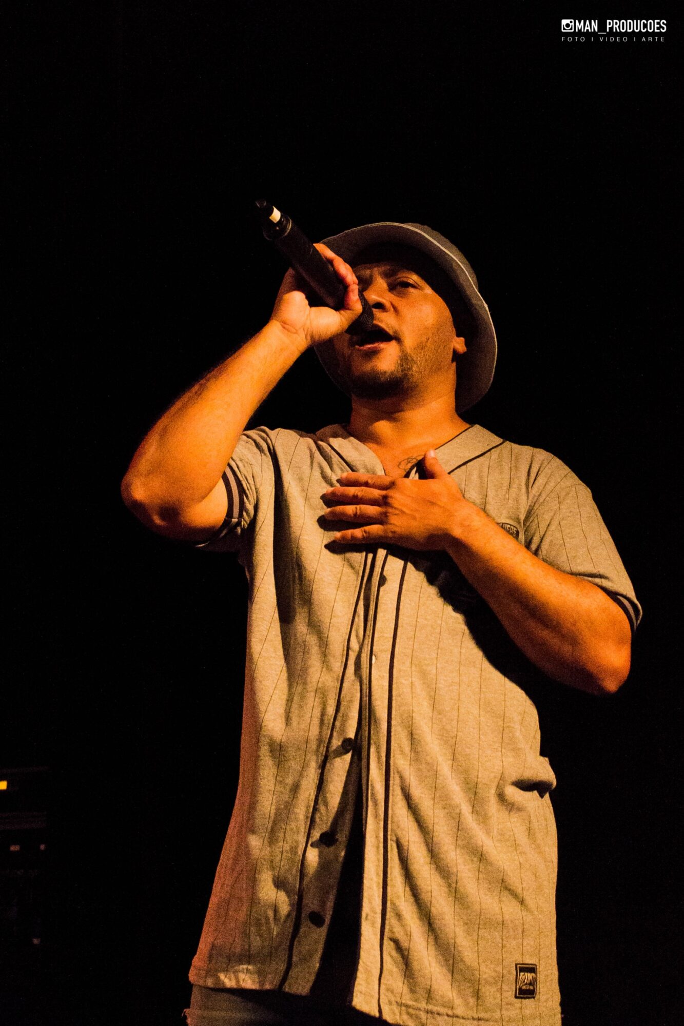 Person singing into microphone with hand on chest, wearing a hat and casual shirt, on stage with dark background.