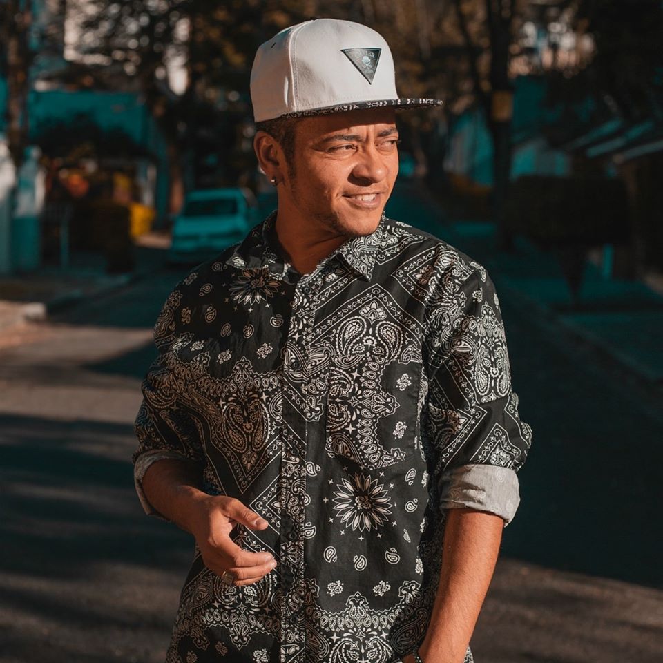 Young man wearing a patterned shirt and white cap, smiling outdoors in sunlight.
