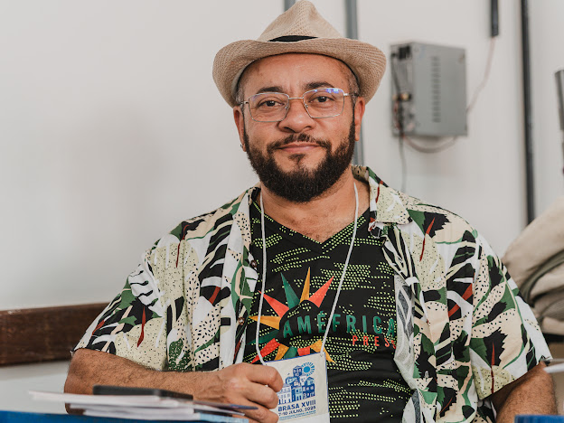 Man with glasses, beard, wearing a hat and patterned shirt, sitting at a table with a badge around his neck.