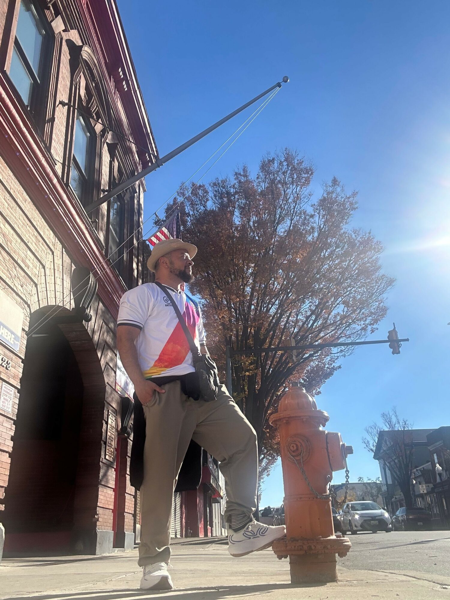 Person standing on sidewalk near brick building, tree with bare branches, blue sky, and utility pole in background.