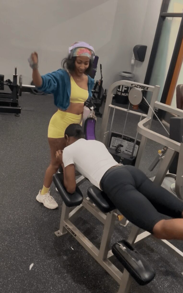 Person lying face down on a workout bench in a gym, with a woman standing nearby, raising her arm. Gym equipment visible.
