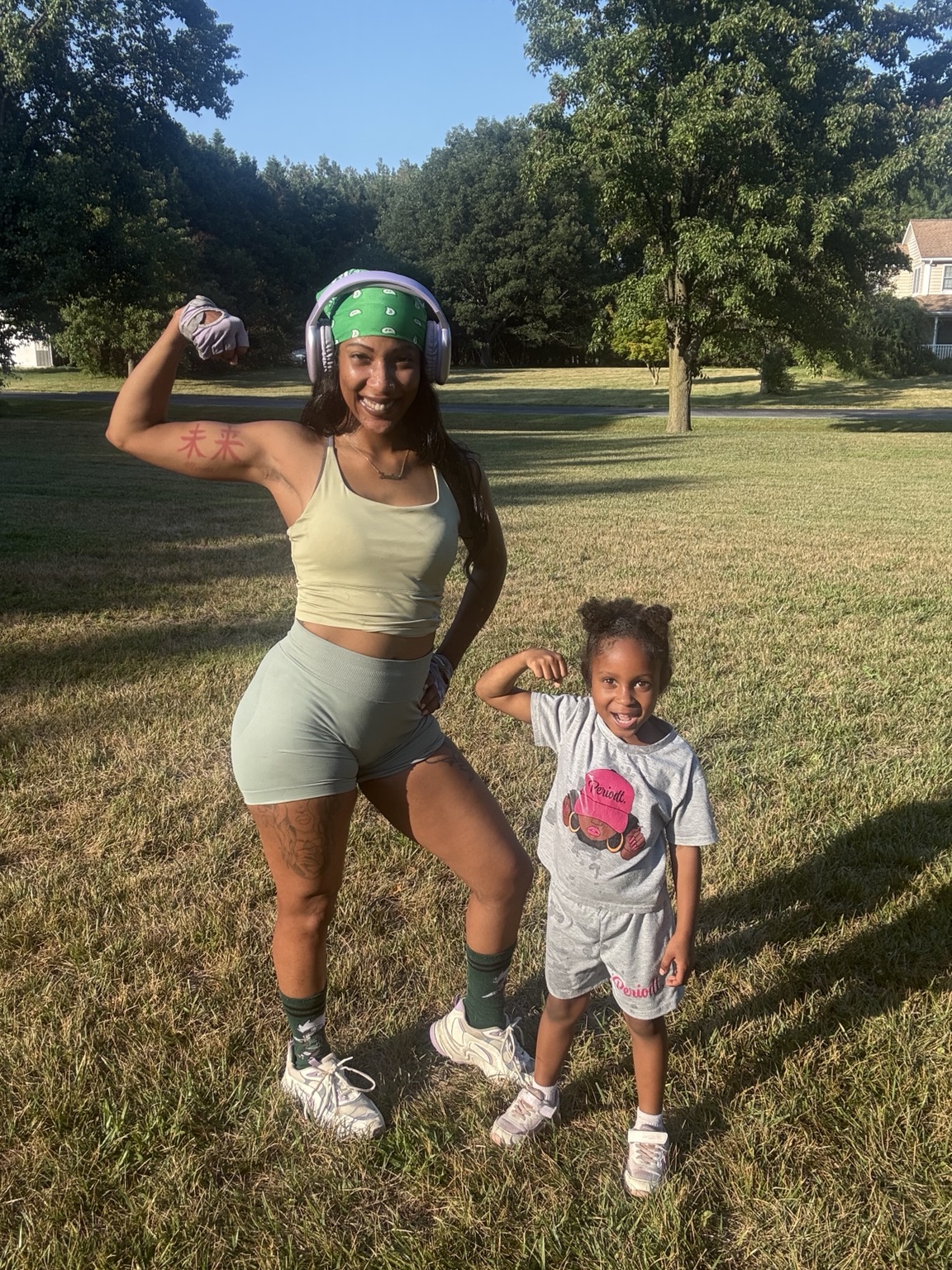 Two girls outdoors, one flexing arm and the other smiling, in a grassy park with trees and a house in background.