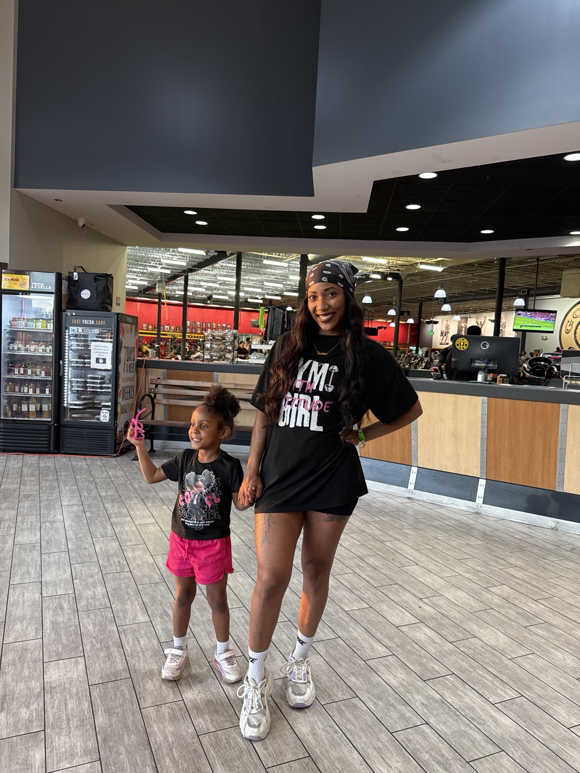 Two girls standing inside a shopping mall, smiling, with a food court and vending machines in the background.