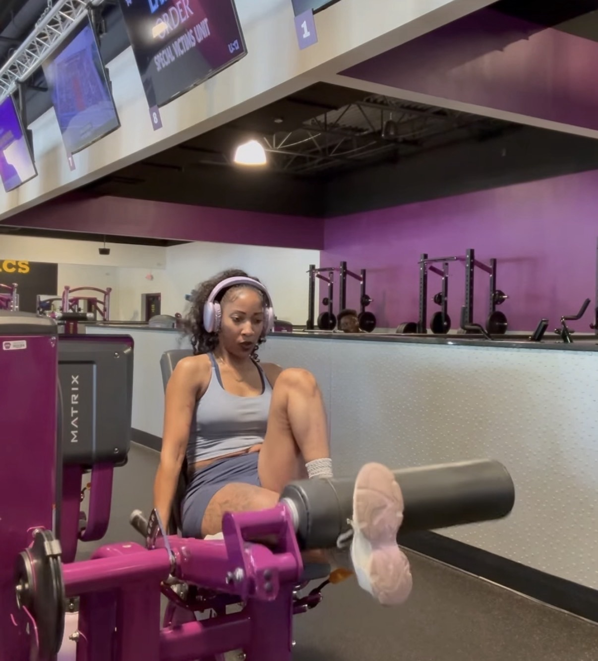 Young woman in workout clothes sitting on a chair in a gym, with one leg extended and foot on a foam roller, headphones on.