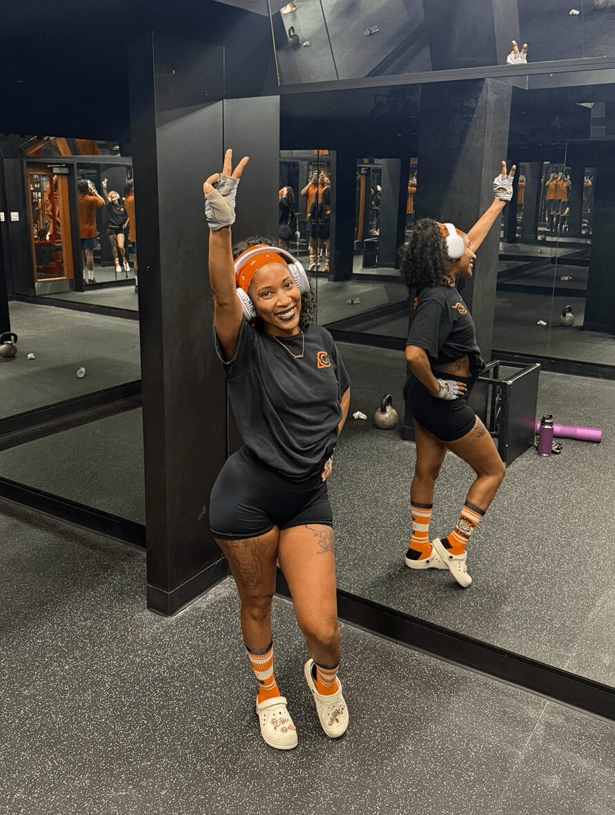 Person in gym wearing black shirt, shorts, orange headband, and striped socks, smiling and making peace signs, with mirror reflection.