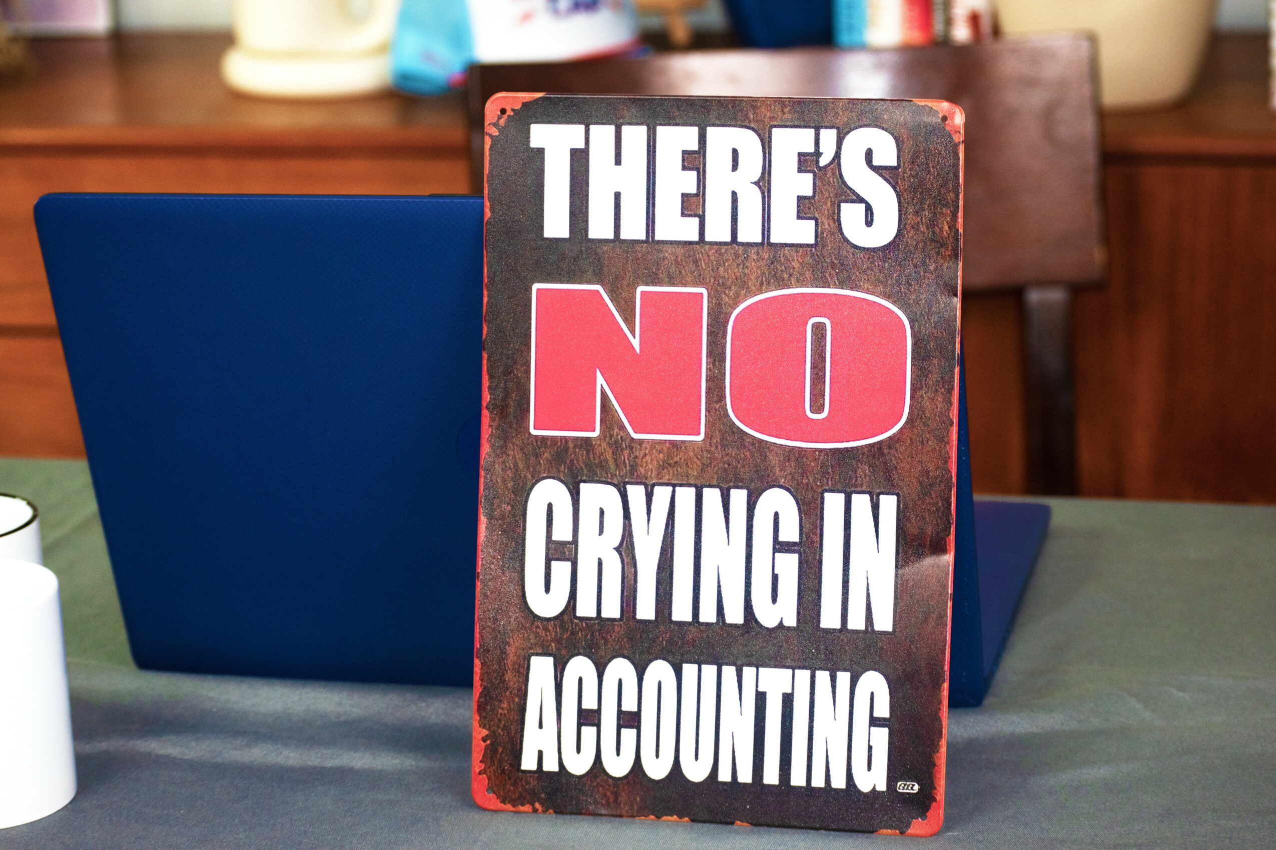 Sign with text: THERE'S NO CRYING IN ACCOUNTING, on a table with a blue chair and background objects