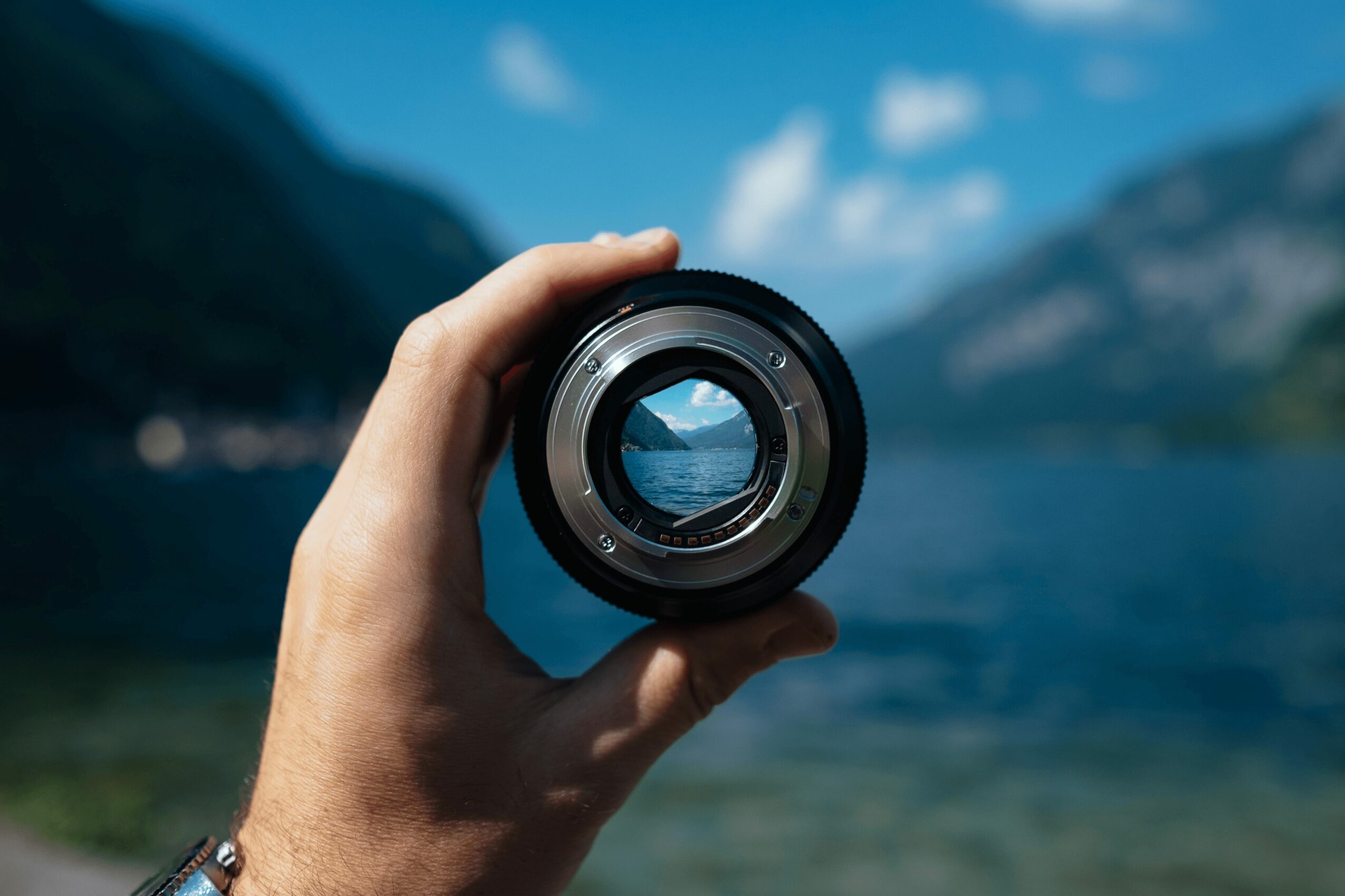 Hand holding a camera lens with a landscape view of mountains and water through it, blue sky with clouds in background.