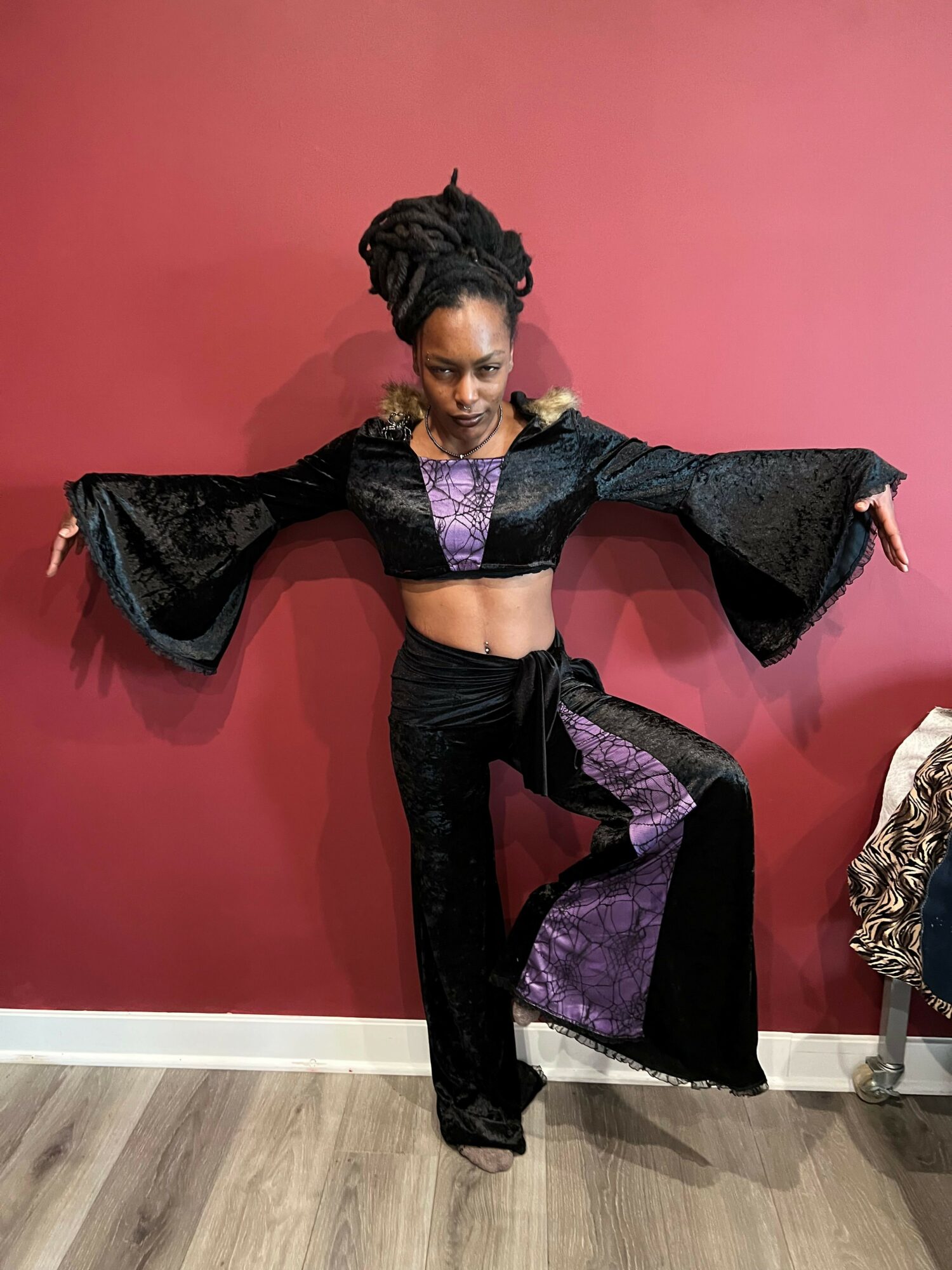 Person lying on a red surface, wearing a black and purple outfit with large bell sleeves, posing with arms and legs extended.