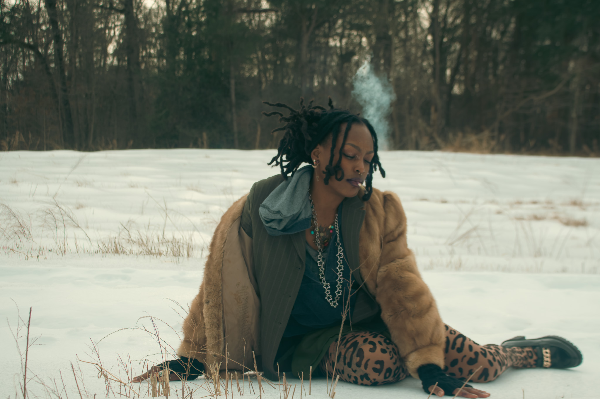 Person with dreadlocks wearing a brown jacket and leopard print pants sitting outdoors in snow, smoking a cigarette.