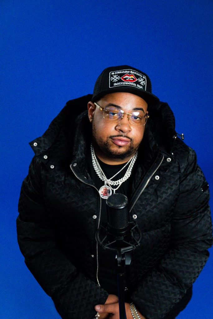 Man wearing glasses, a black cap, and layered necklaces, standing in front of a blue background.