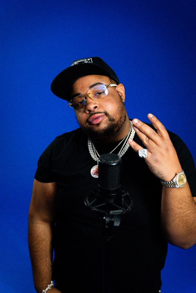 Man wearing glasses, a black cap, and jewelry, standing in front of a blue background, making a hand gesture.