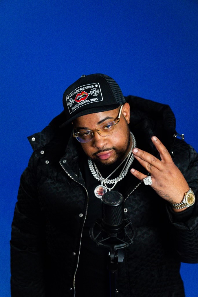 Man wearing glasses, a black cap, and jewelry, posing with hand gesture in front of a blue background.