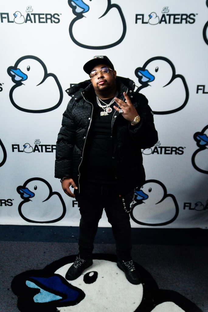 Man in black clothing and jewelry standing in front of a duck-themed backdrop, making a hand gesture.