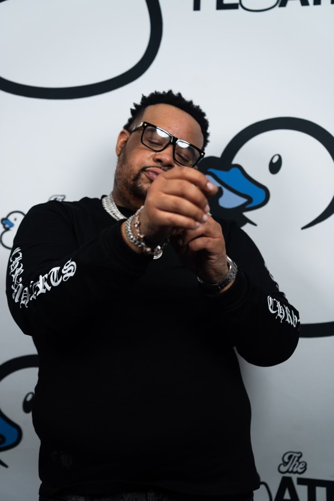 Man with glasses and jewelry, wearing a black shirt, standing in front of a white background with cartoon duck illustrations.