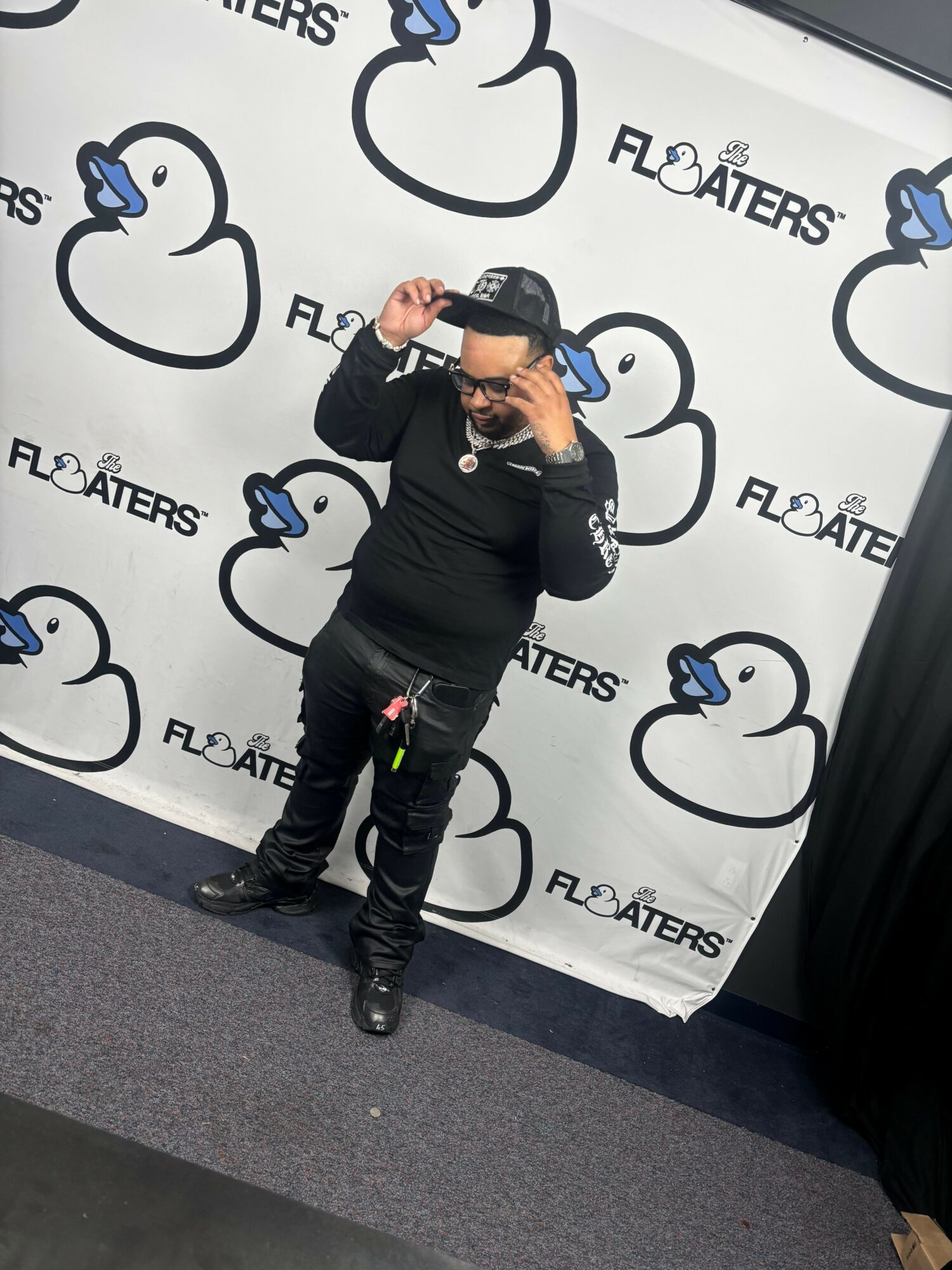 Person standing against a backdrop with a cloud logo and text, adjusting their cap, wearing black clothing and boots.