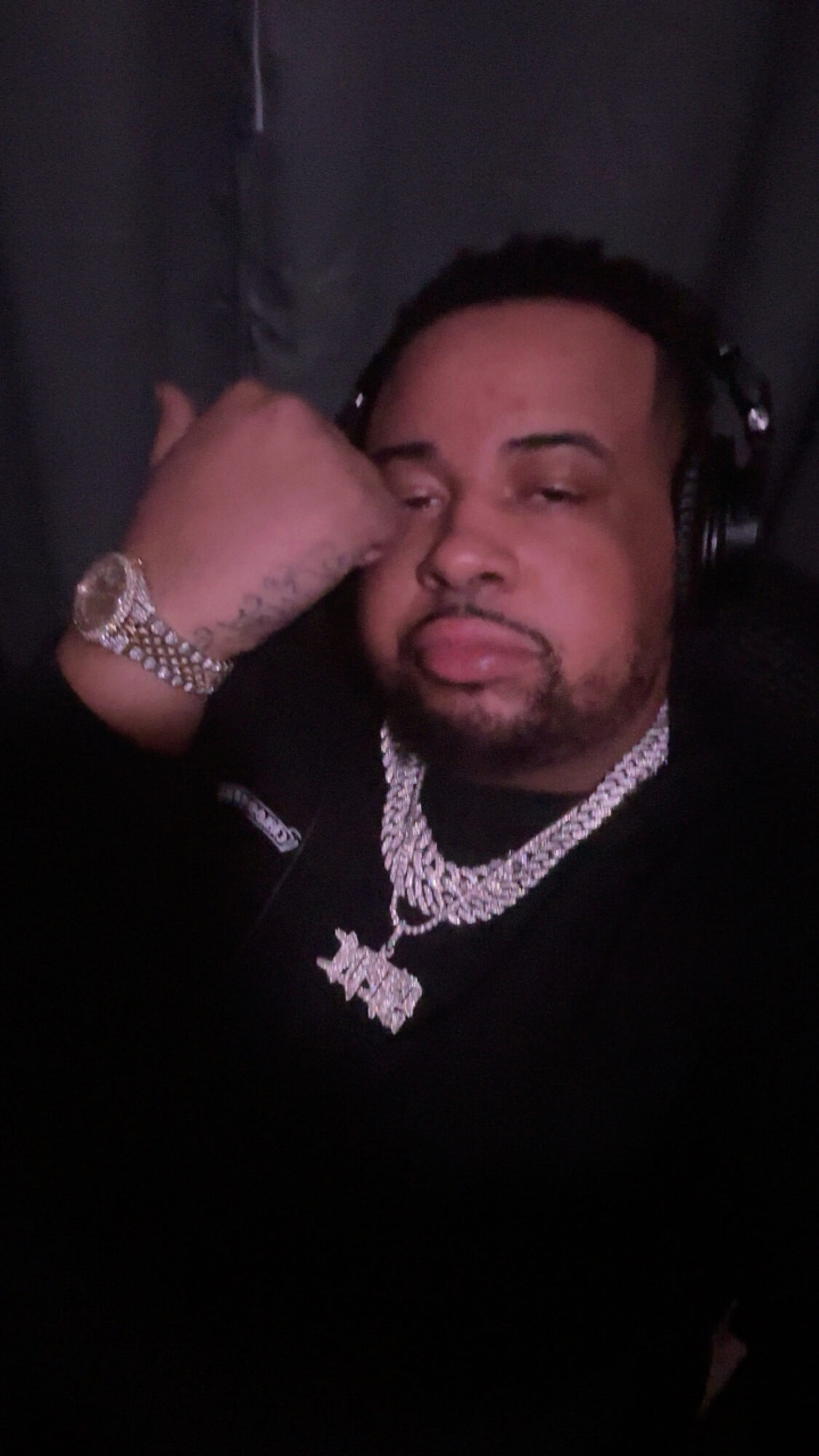 Person with headphones, wearing jewelry, resting head on hand, looking at camera, dark background