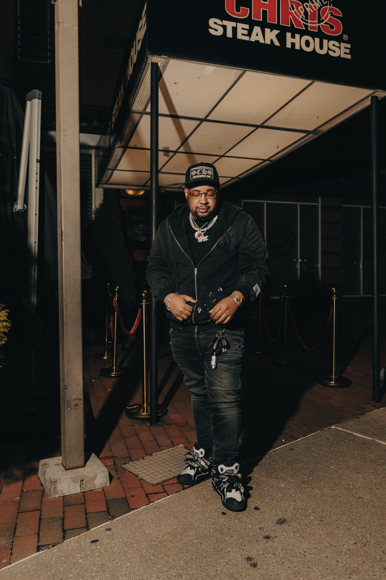 Person standing outside a restaurant at night, wearing a black hoodie, cap, and sneakers, with a sign reading 'Steak House' above.