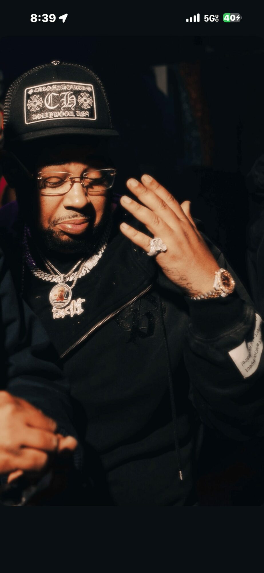 Person wearing a cap, glasses, and jewelry, touching their face with one hand in a dark setting.