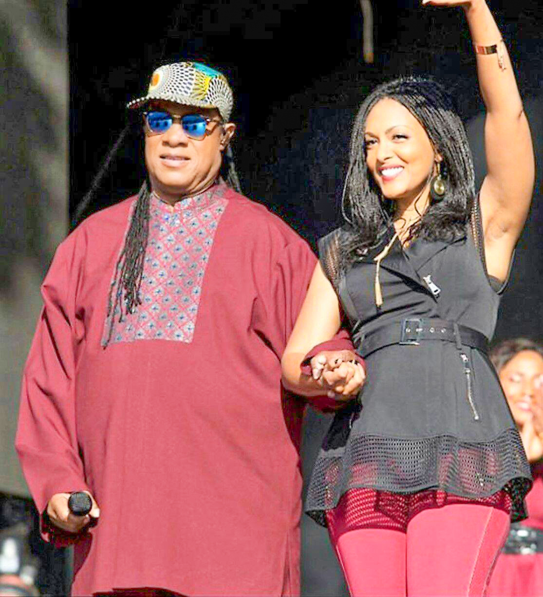 Two people on stage, one in a red outfit and the other in black and red, smiling and holding hands.