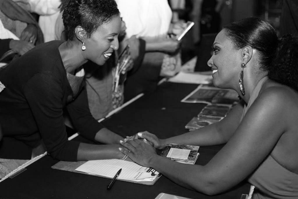 Two women smiling and holding hands across a table, engaging in conversation, with notebooks and pens nearby.