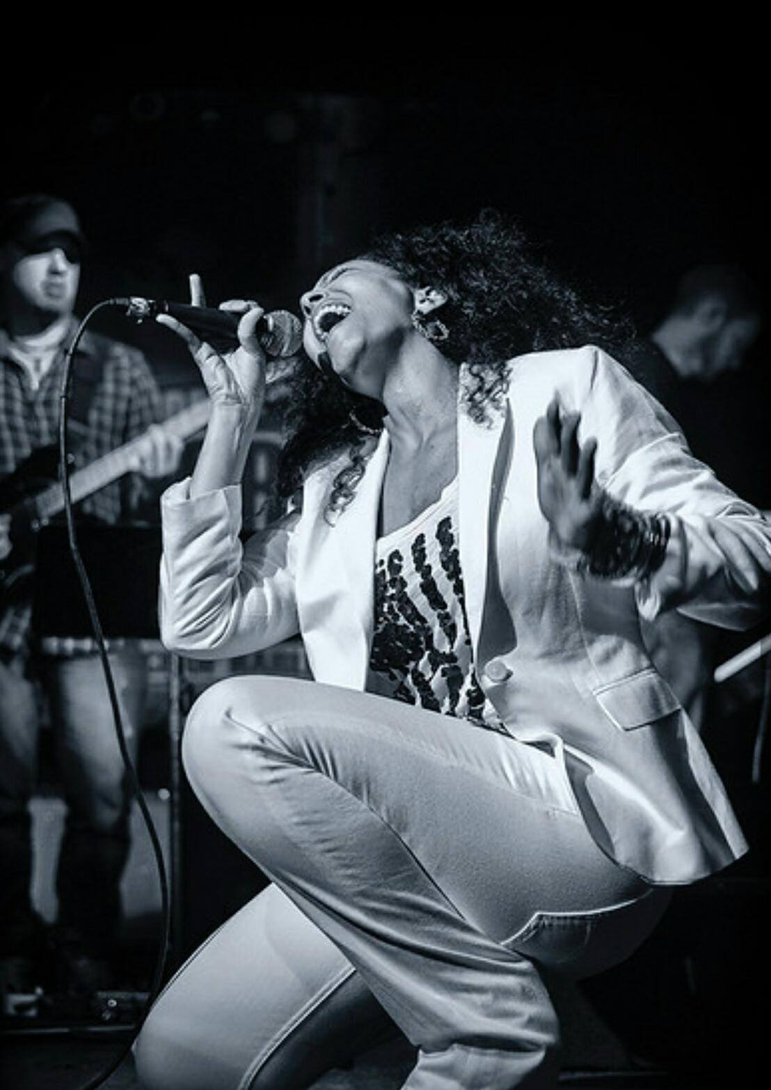 A woman singing passionately into a microphone on stage, with musicians in the background, in black and white.