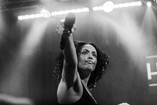 Woman with curly hair holding a microphone on stage, illuminated by bright lights, looking upward.