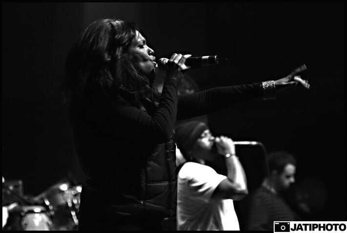 Three women singing into microphones on stage, with one woman in the foreground and two in the background, in black and white.