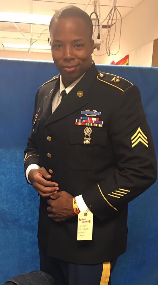 Young man in military uniform standing indoors, smiling, with decorations and a blue background.