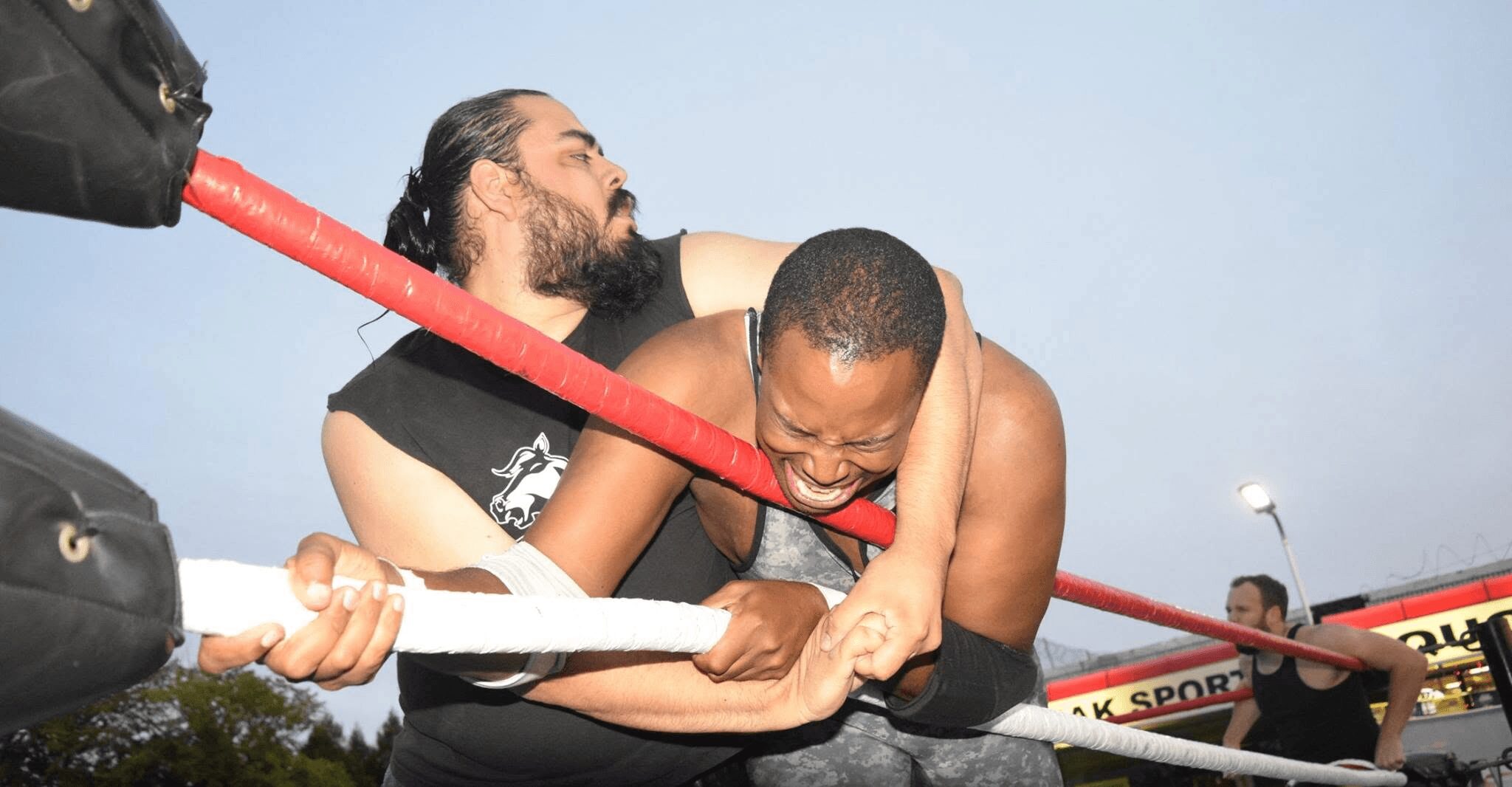 Two wrestlers in a ring, one holding the other in a headlock, with a red rope in the background.