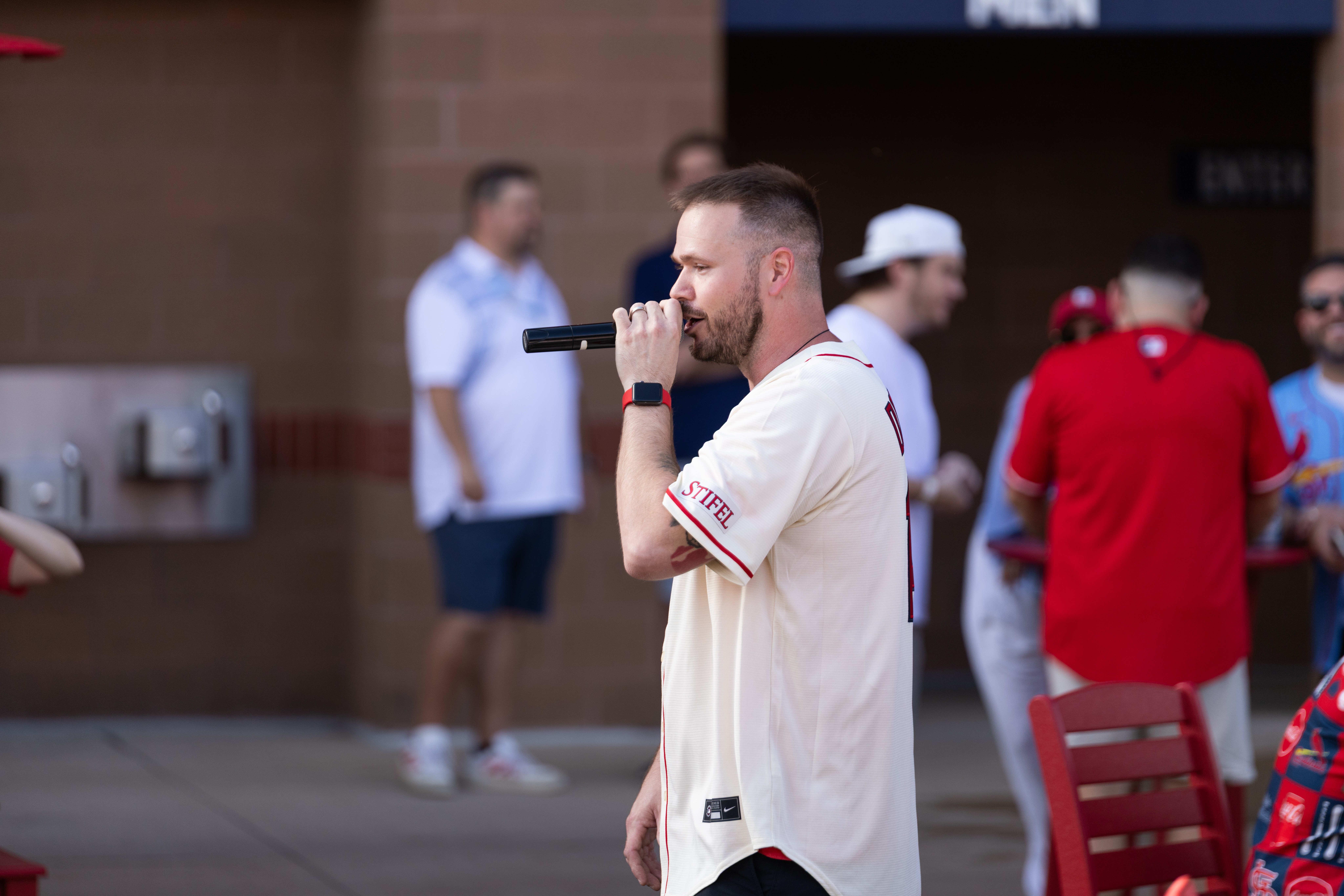 Man holding a microphone and speaking outdoors, with people in the background wearing casual clothing.