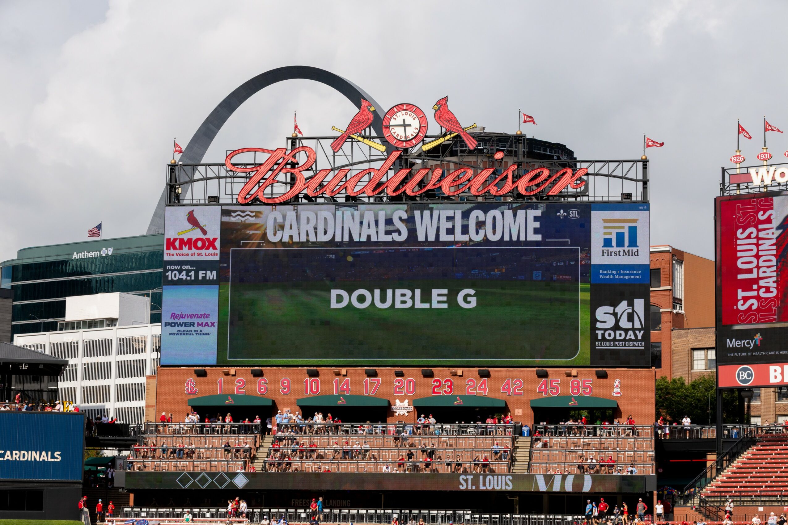 St. Louis Cardinals stadium with scoreboard displaying game information, fans in stands, and city skyline in background.