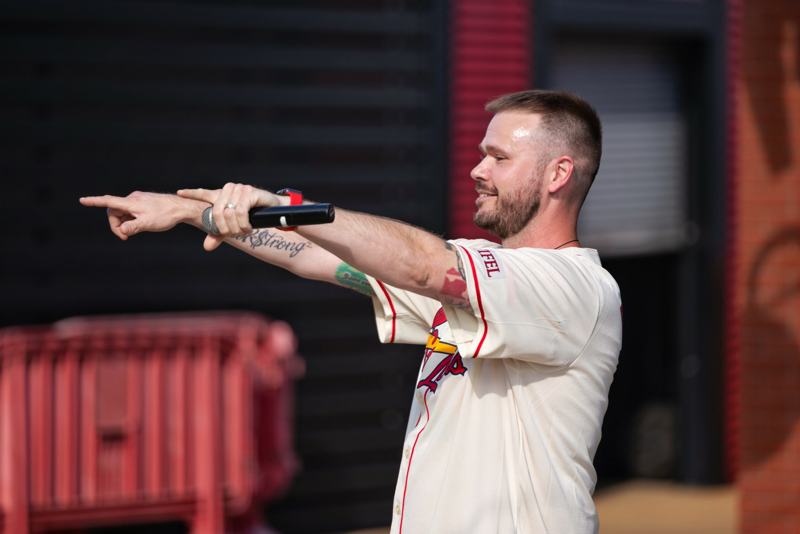 Man with a beard and tattooed arm pointing with both hands, wearing a white sports jersey, in front of a red and black background.