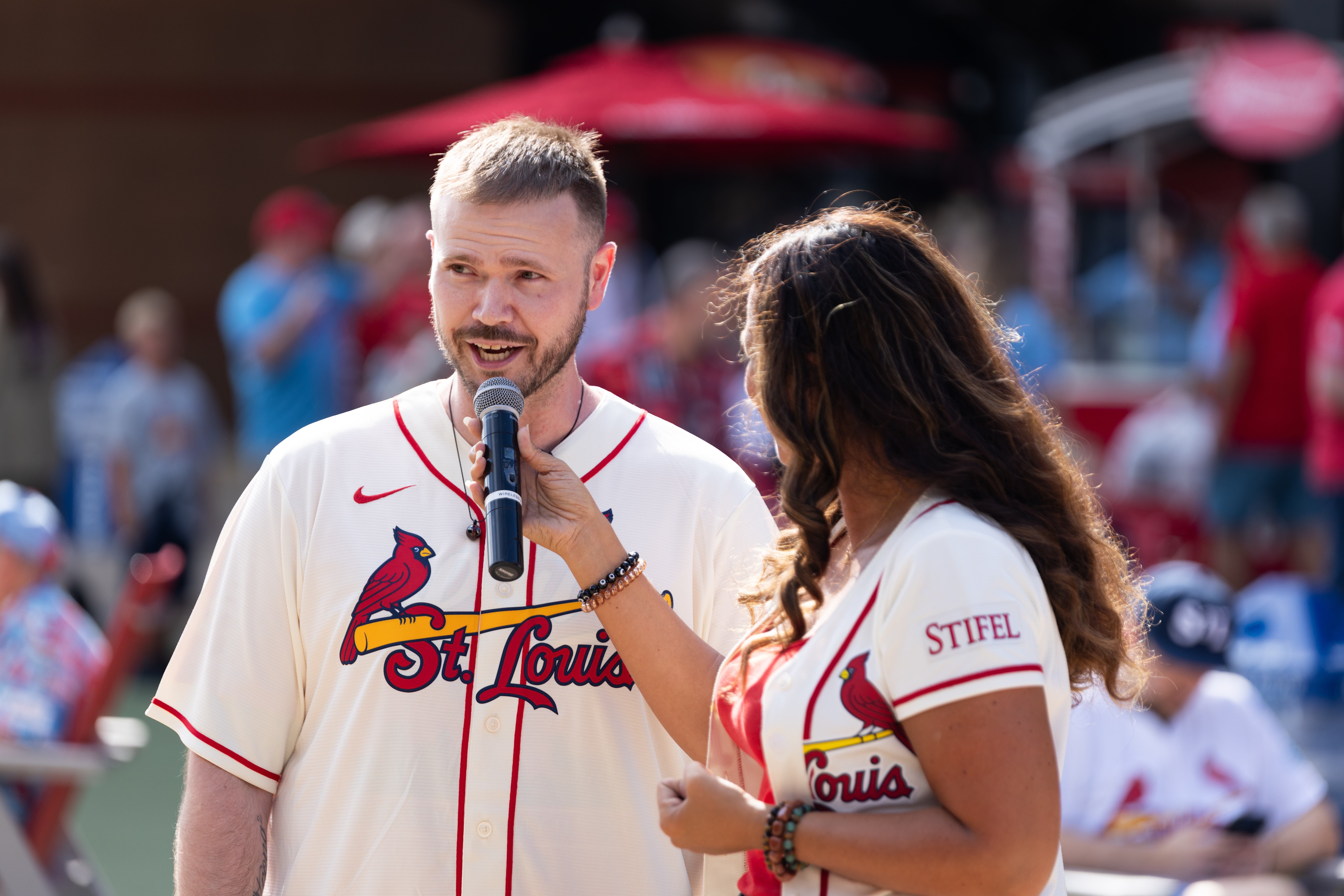 A man with a beard and a woman with long dark hair are at a sports event, with the woman holding a microphone to the man.