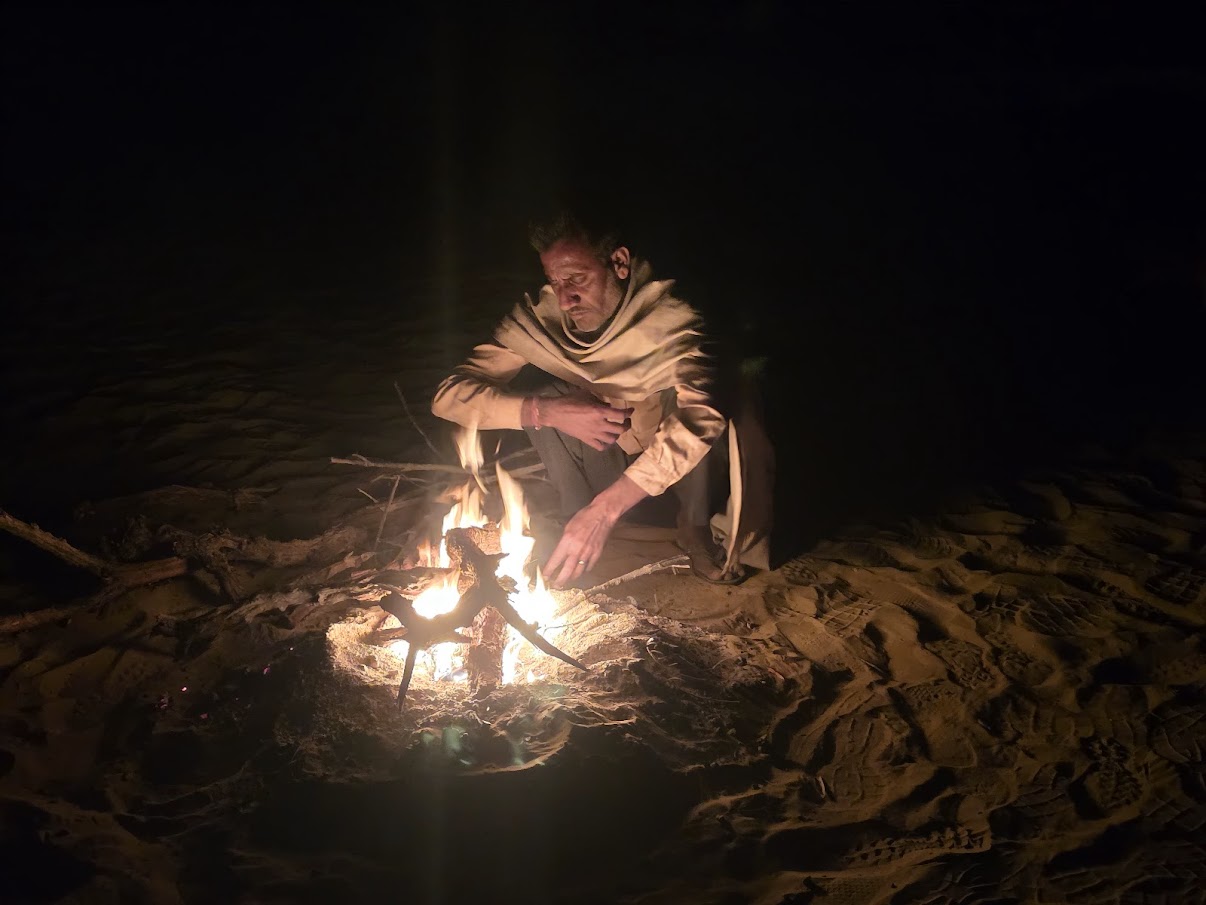 Person sitting near a small campfire on sand at night, with darkness surrounding.